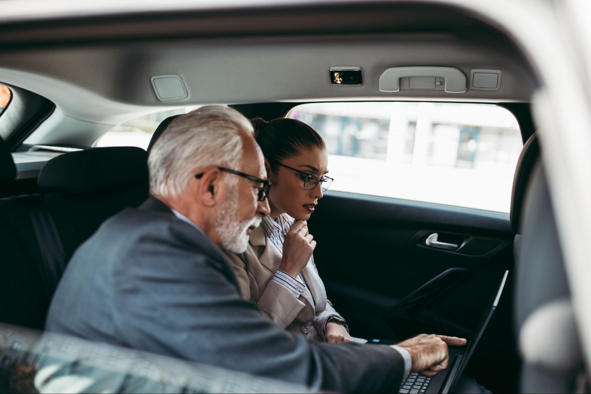 An older gentleman describes what he sees on a laptop screen to a younger executive while carpooling in a vehicle.