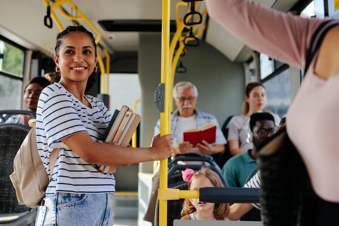 Young girl holding books rides a filled bus while looking happy.