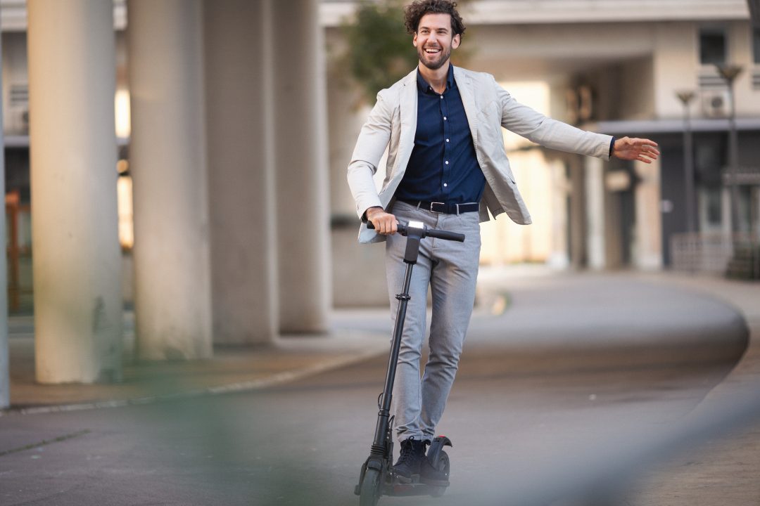 A businessman with a white jacket and grey slacks rides an electric scooter to his destination.